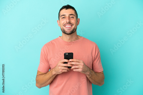 Young handsome caucasian man isolated on blue background looking at the camera and smiling while using the mobile