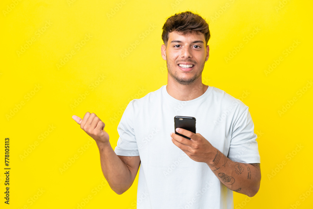 Young caucasian handsome man isolated on yellow background using mobile phone and pointing to the lateral Young caucasian handsome man isolated on yellow background using mobile phone and pointing to the lateral