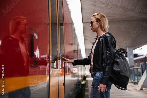 Young blond woman in jeans, shirt and leather jacket wearing bag and sunglass, presses door button of modern speed train to board on train station platform. Travel and transportation.