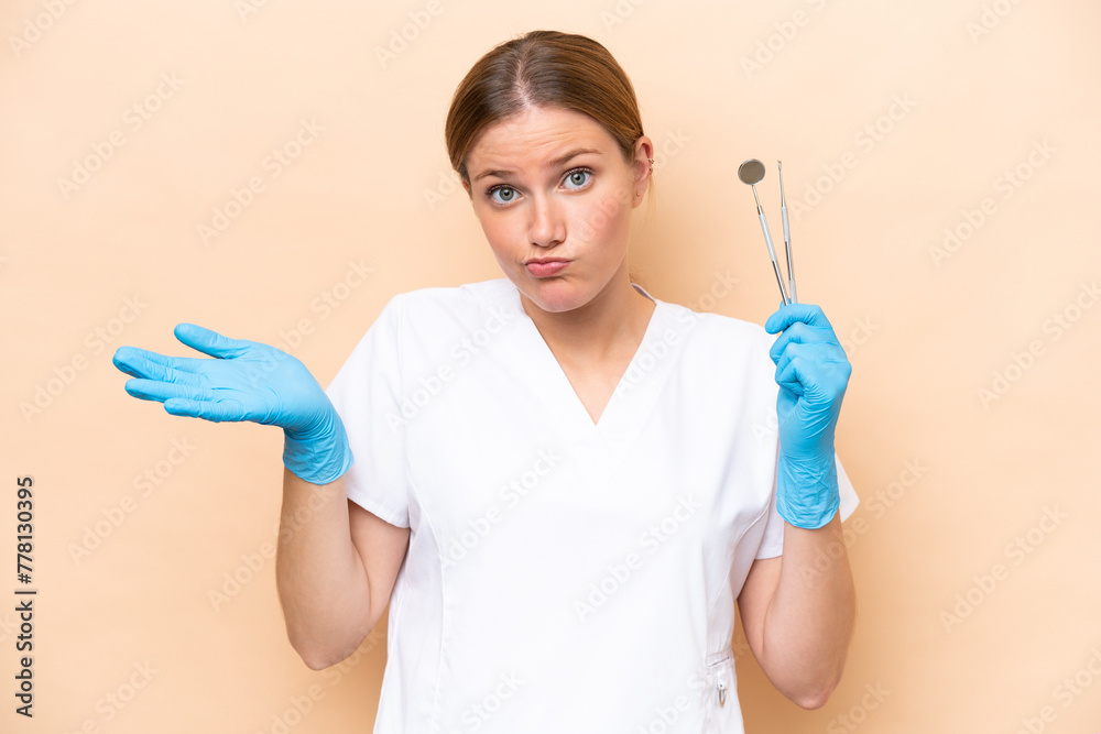Dentist caucasian woman holding tools isolated on beige background having doubts while raising hands Dentist caucasian woman holding tools isolated on beige background having doubts while raising hands