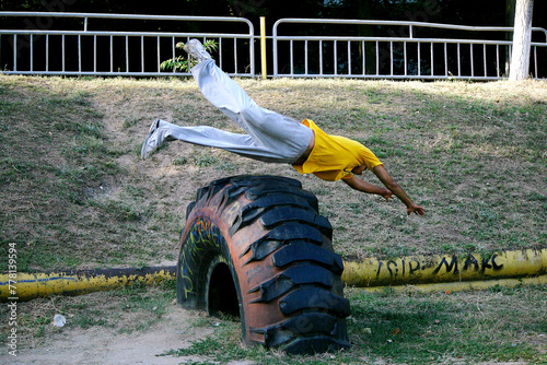A young guy in a yellow T-shirt performs an acrobatic trick in parkour style at the stadium through a huge tire photo