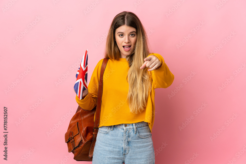 Young blonde woman holding an United Kingdom flag isolated on white background surprised and pointing front Young blonde woman holding an United Kingdom flag isolated on white background surprised and pointing front
