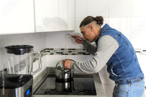 Caucasian man cooking in the kitchen with a pot and a ladle by hand in the natural light, on a ceramic hob, is tasting food, side view. Concept of male doing housework