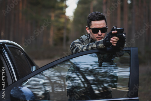 A male fbi agent in sunglasses aims a pistol from behind a car door in the forest. photo