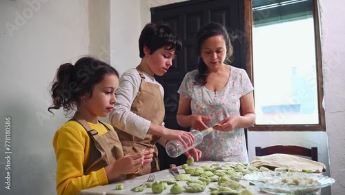 Lovely kids in beige chef apron, standing at floured kitchen table, helping their mother to stuff and sculpt dumplings. Mom, daughter and son cooking together varennyky according to traditional recipe