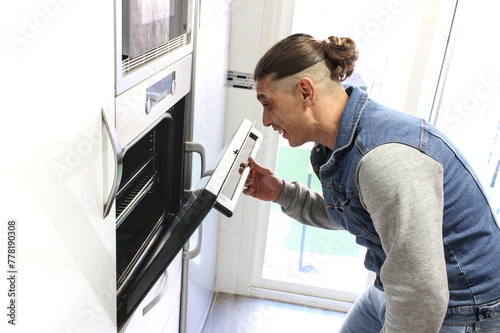 Caucasian man opening oven and looking inside smiling, inside house, natural lighting, side view. Concept of male doing housework
