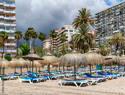 Sandy beach with sun loungers and straw umbrellas against the backdrop of a stormy sky before the rain in Marbella, Spain.