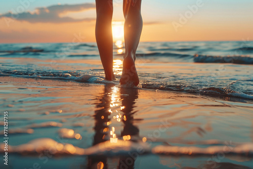 Close-up photo of woman's legs from behind as she walks along sandy beach that is washed by sea water against sunset background. Beautiful summer vacation background