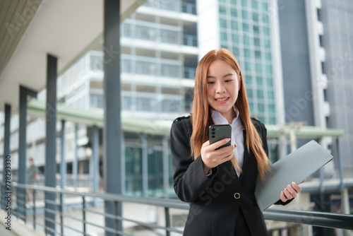 A young Asian businesswoman wearing a suit holding files standing in a big city waling on the street. Young Asian businesswomen using smartphone texting to contact clients.