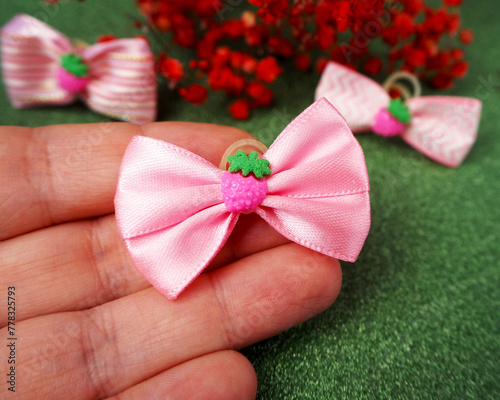 On a green background, in your hand lies a small pink bow with a strawberry in the middle. side view . accessories for long haired dogs. top notes