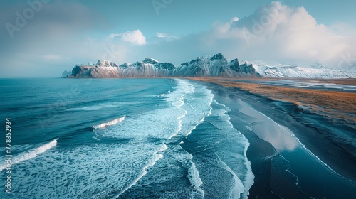 Drone view of rocky mountains covered in snow near waving seawater along black sand beach in Iceland, eystrahorn, krossanesfjall, iceland, against cloudy skies photo