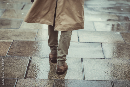 A man wearing a brown coat and brown shoes walks on a wet sidewalk