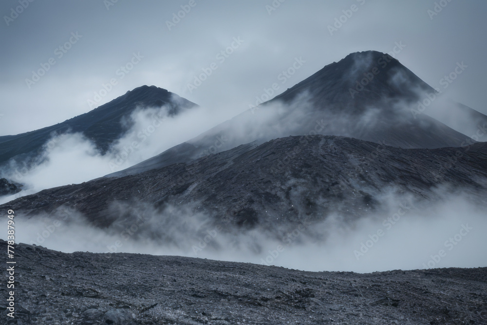 Snowy volcanic peaks pierce a cloudy sky, while a lower mountain disappears into a misty landscape