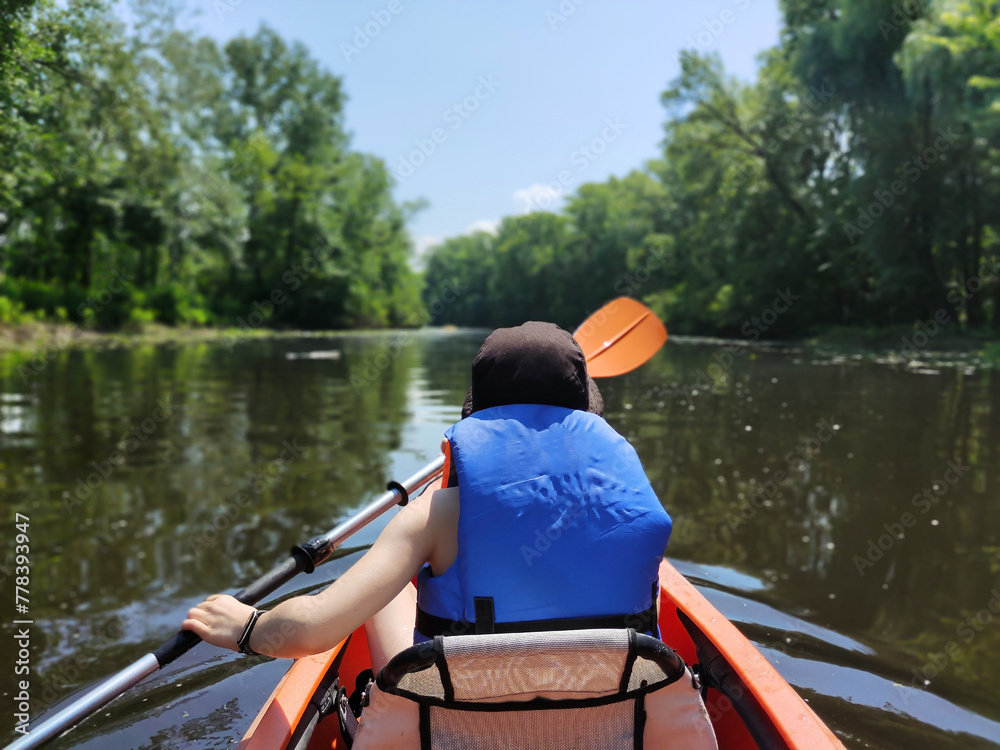 A teenage boy is swimming in a life jacket in a kayak on the river against the background of trees. Rear view