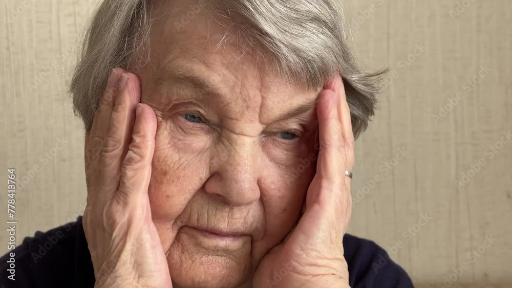 A close-up of the head of serious mature old woman sitting at home on the couch and holding her hands to her temple. The excitement, the anxiety, the feeling of pain in her eyes A close-up of the head of serious mature old woman sitting at home on the couch and holding her hands to her temple. The excitement, the anxiety, the feeling of pain in her eyes