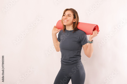 Young smiling woman in sportswear is holding a pink fitness mat over her shoulders.