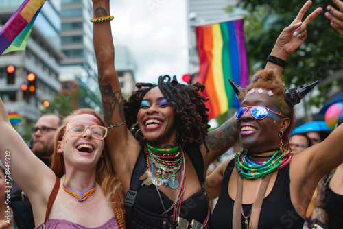 Three women are smiling and holding up rainbow flags. They are dressed in colorful clothing and appear to be celebrating something