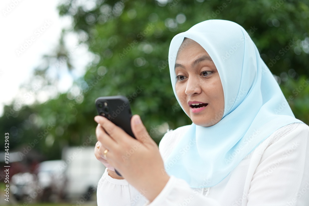 Asian woman in blue hijab is holding a cell phone with her facial expression while outdoors in the bright sun Asian woman in blue hijab is holding a cell phone with her facial expression while outdoors in the bright sun