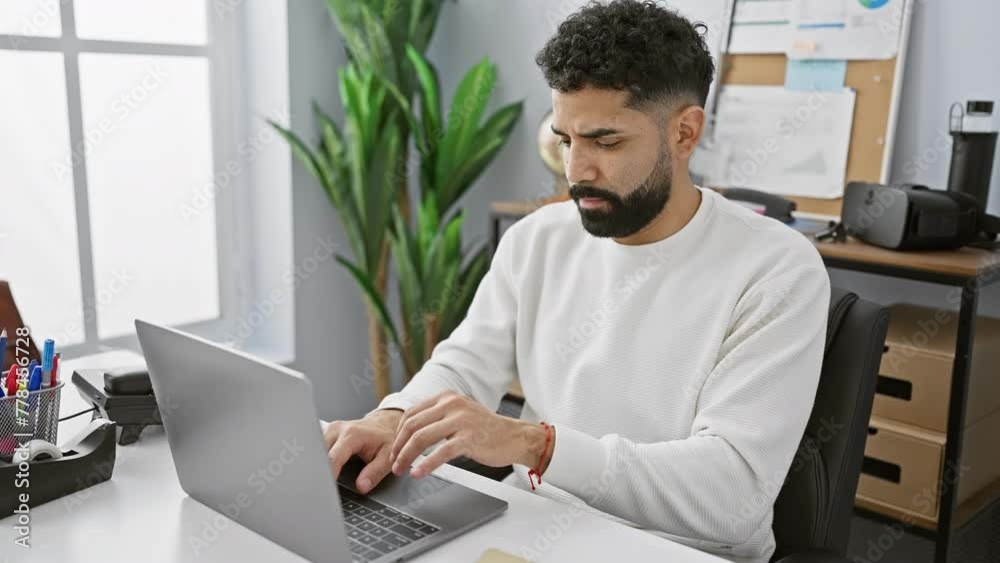 A young bearded man experiencing shoulder pain while working on a laptop in an office setting. A young bearded man experiencing shoulder pain while working on a laptop in an office setting.