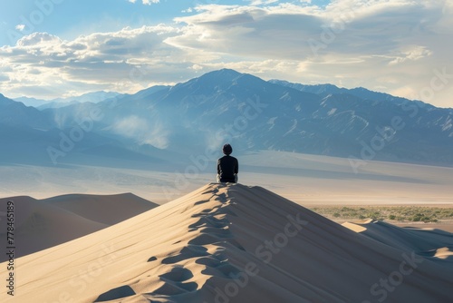 A solitary person sits on the edge of a high desert dune, overlooking a vast mountain landscape. Solitary Figure Sitting on Desert Dune Edge