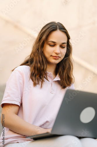 Young smiling female student is sitting on some stairs working on her laptop outdoors.