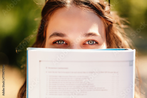 Cheerful young woman is hiding behind a book showing only her blue eyes. Outdoors shot.