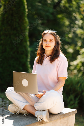 Young concentrated woman is sitting in park using laptop for her studies on a sunny day.