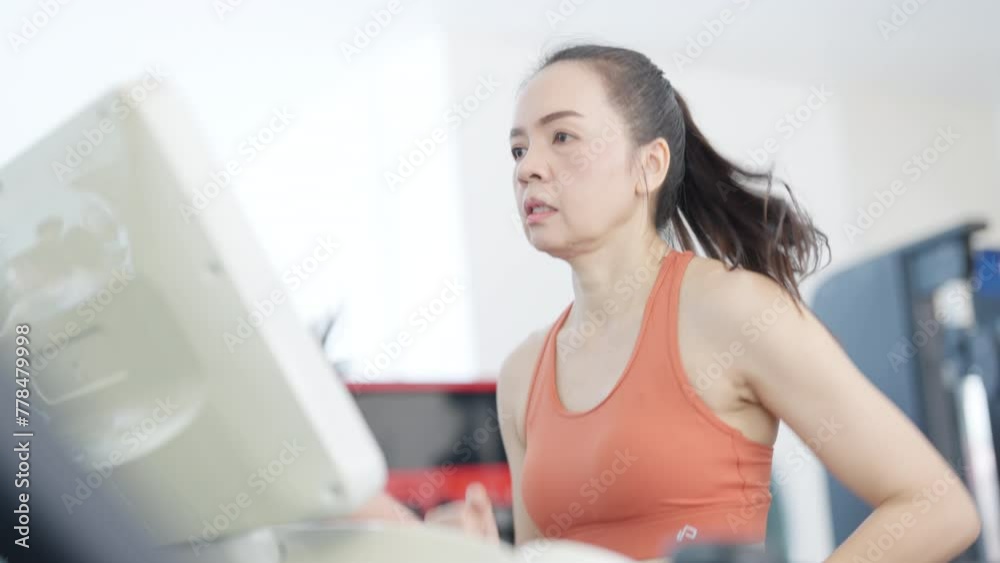 A woman is running on a treadmill in a gym. She is wearing an orange tank top and is looking at a computer screen A woman is running on a treadmill in a gym. She is wearing an orange tank top and is looking at a computer screen