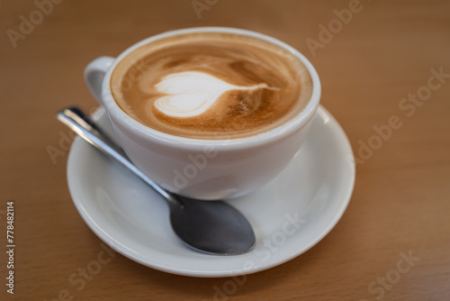 Shallow focus on the bubbles in the froth of a flat white coffee in a white cup and saucer and a metal teaspoon on a wooden table.