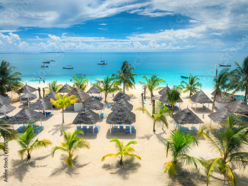 Aerial view of white sandy beach with palm trees, umbrellas, yachts, boats, blue ocean, sky with clouds at sunset. Summer vacation in Kendwa, Zanzibar island. Tropical landscape. Clear sea. Top view