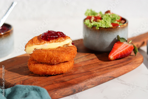 A portion of fresh made fried camembert cheese with cranberry sauce on top and a side of fresh salad on wooden plate