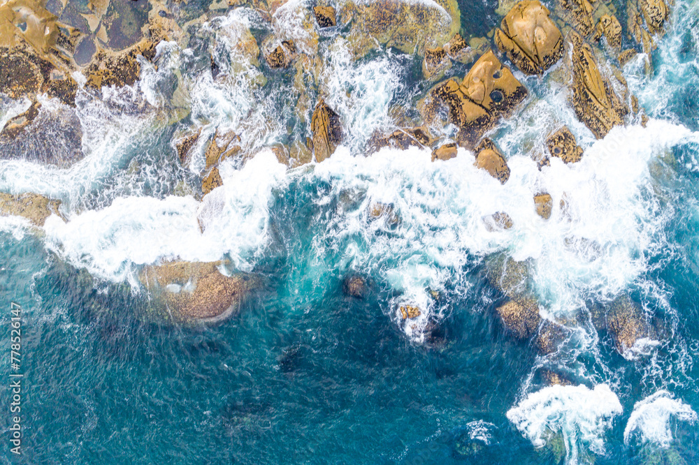 Turquoise sea surface and white sea foam and waves splashing the rocks on the shore. Top down view with drone