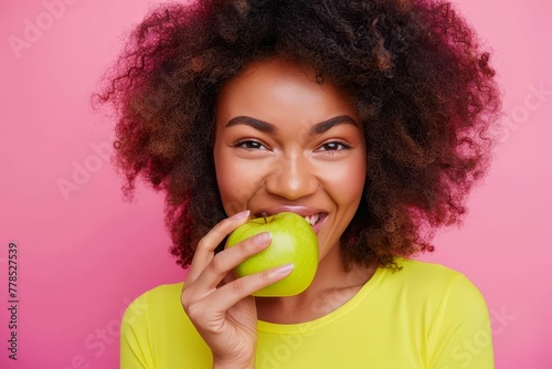 Beautiful smiling woman in bright yellow top biting crisp green apple, isolated on pink background, concept of happiness and healthy eating photo