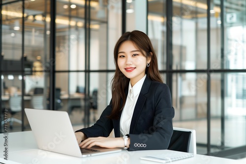 A young asian woman in a suit sitting at a desk with a laptop in office