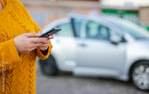 A woman is holding a cell phone while standing next to a silver car. The scene suggests that she might be checking her phone for directions or information about the car.