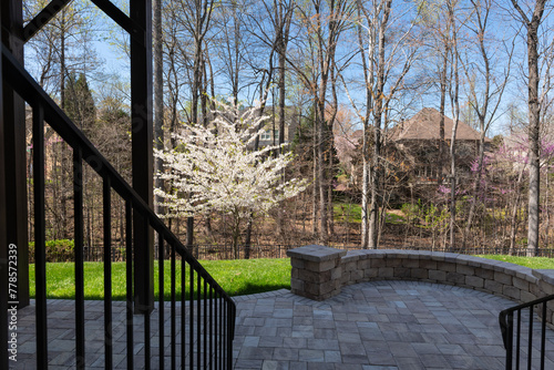 Picturesque backyard view in spring season with patio pavers and stone wall, blooming white cherry tree, and spring colored woods in the background.