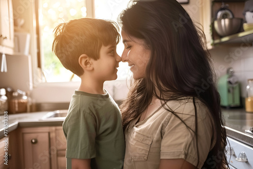 A mother and son share a tender moment, touching noses and smiling in a sunlit kitchen. photo