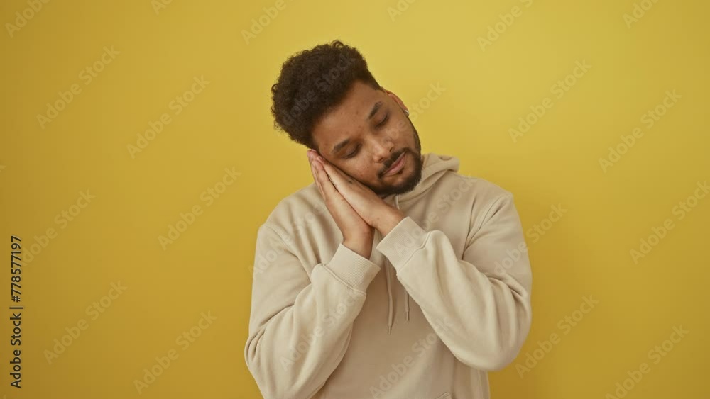 Happy african american man napping in hoodie, posed with hands together tiredly, smiling as he dreams, isolated on a yellow background. Happy african american man napping in hoodie, posed with hands together tiredly, smiling as he dreams, isolated on a yellow background.