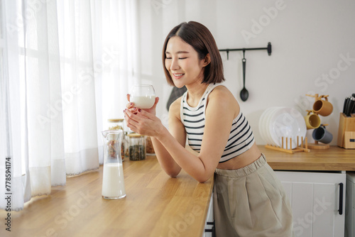 Smiling woman in a casual outfit enjoys a fresh glass of milk in her bright kitchen, starting her day on a healthy note