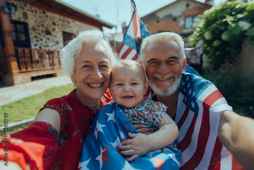 Happy loving family with seniors and baby holding American USA Flag draped over shoulders on backyard of typical American house at sunny day. Family celebrates 4 July Independence Day