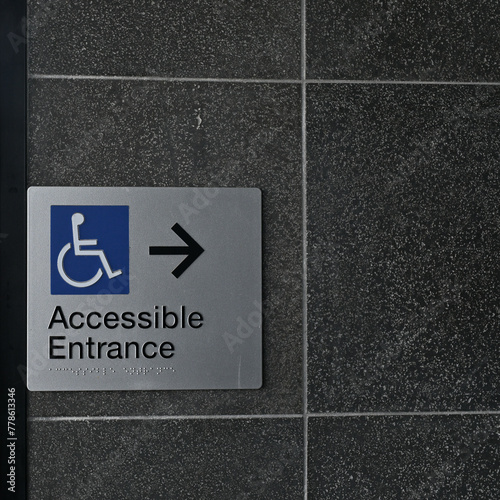 Rectangular accessible entrance sign, featuring blue wheelchair disability icon, black text, braille, and a black arrow on a dark wall