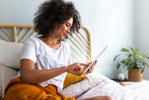 Young African American woman using tablet lying down relaxing on bed at home cozy bedroom.
