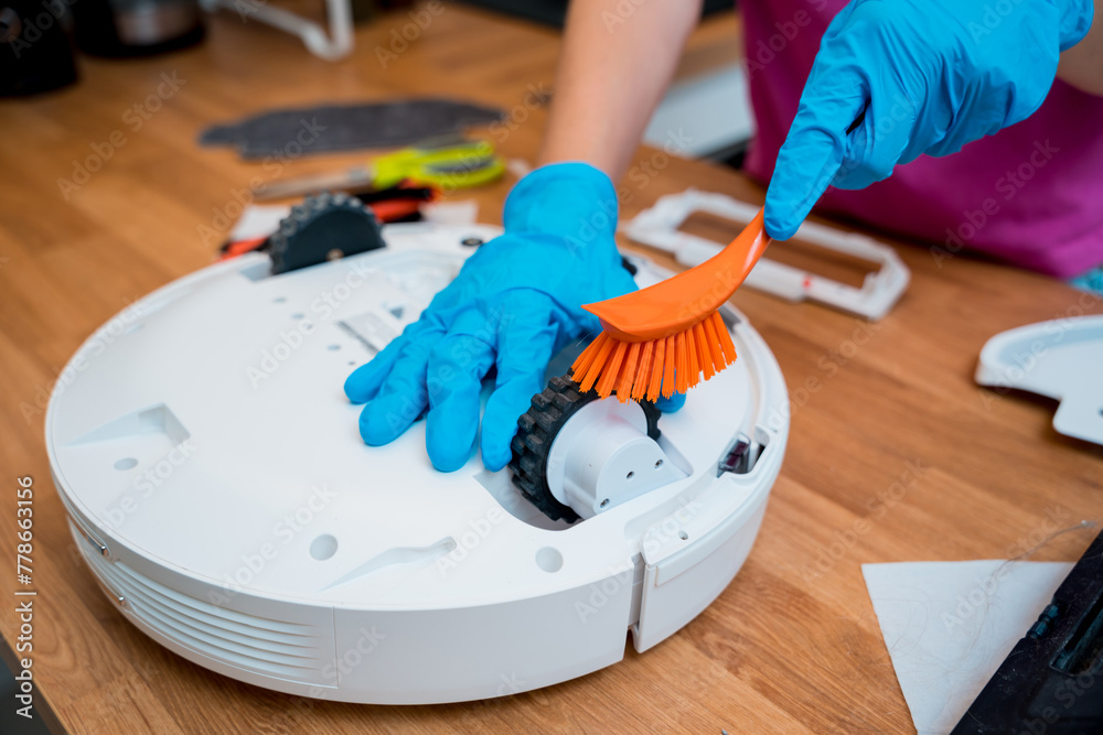 A young woman cleans a robot vacuum cleaner from dirt after cleaning. - obrazy, fototapety, plakaty 