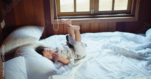 A Asian child girl wearing pajamas is laying resting comfortably on white bed while lying amidst rumpled white sheets in a wooden-paneled room with windows letting in natural light in the morning