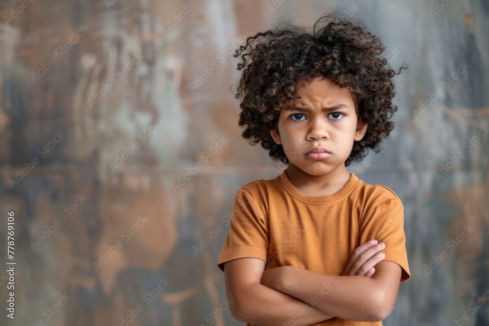 A young boy with voluminous curly hair stands scowling with his arms crossed A young boy with voluminous curly hair stands scowling with his arms crossed