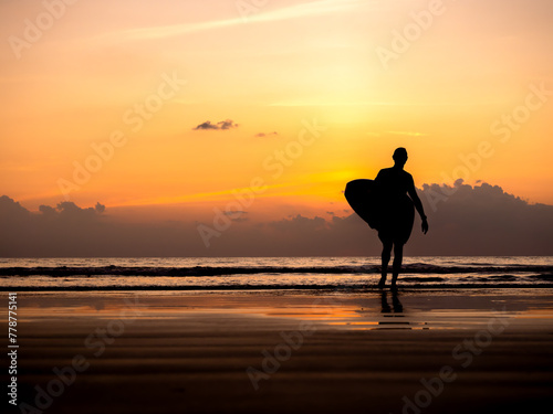 Man silhouette holding surfing board, walking on dark seashore with copy space. Surfer standing on sunset sky over the sea water background. Summer sand beach in evening time. dusk seascape scene.