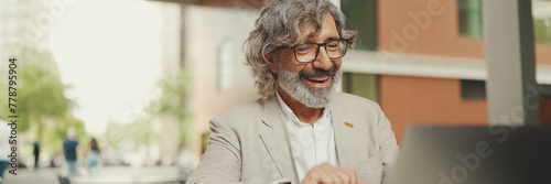 Mature businessman with beard in eyeglasses wearing gray jacket sits drinks coffee in cafe. Panorama of middle aged manager successful man working on laptop pc computer sit at cafe outdoors