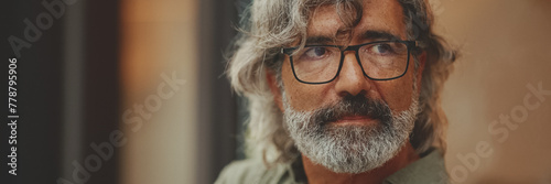 Panorama of thoughtful middle-aged man with gray hair and beard, wearing casual clothes, sits in street cafe. Mature gentleman in eyeglasses