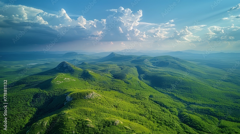   An aerial view of a green mountain range under a cloudy blue sky with a sunbeam illuminating the center of the image