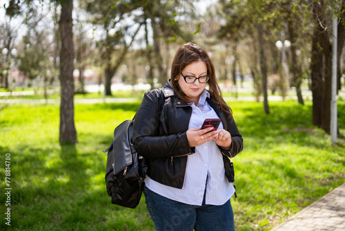 A chubby woman in a short leather bag and casual jeans walks through the park with a phone in her hands. Curvy girl looking at the phone